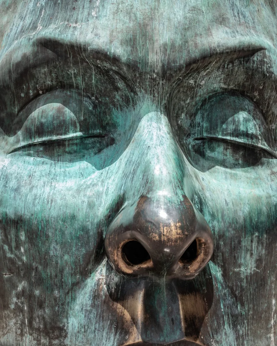Detalle de un rostro de bronce con pátina verdosa y desgastes que dejan ver el metal